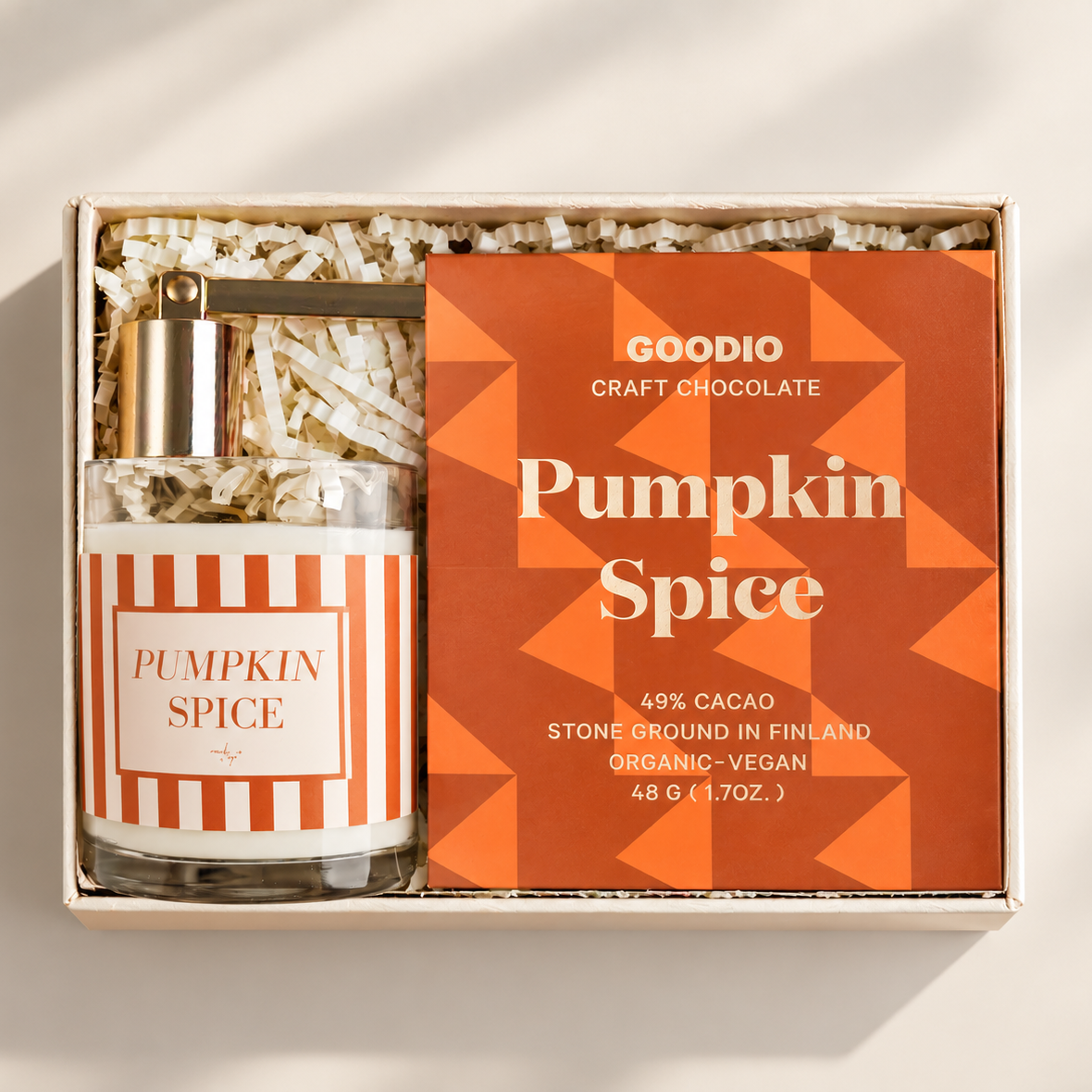 A gift set in a light wood box with shredded paper filler, featuring a clear glass bottle labeled 'Pumpkin Spice' with a gold cap and an orange geometric box labeled 'GOODIO Craft Chocolate Pumpkin Spice'.