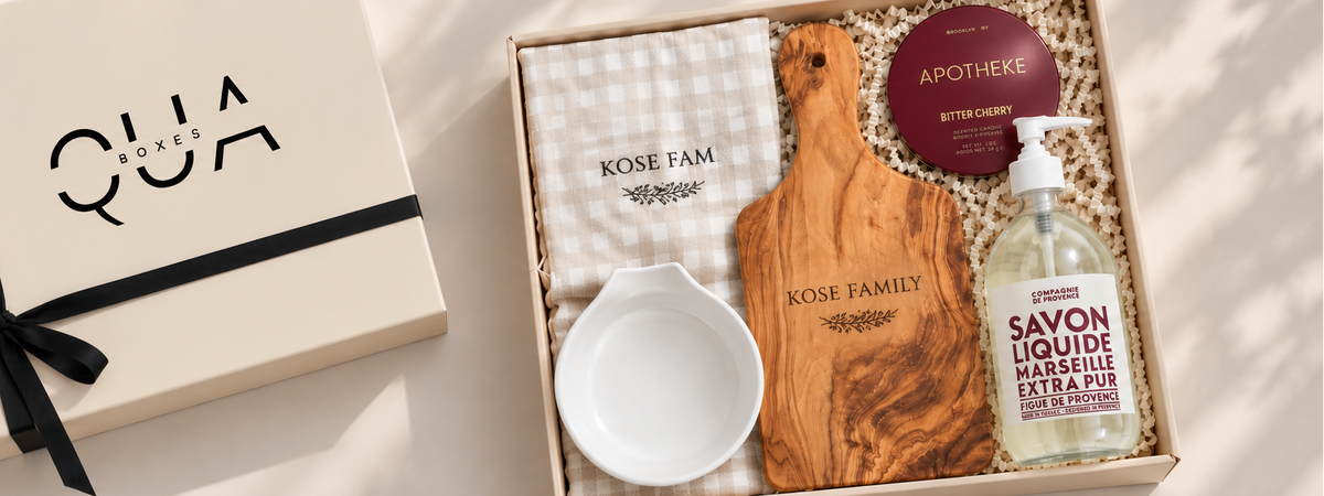 A curated gift set from QUA, featuring a cream-colored box with a black ribbon on the left, and an open gift box on the right containing a wooden cutting board, a white ceramic bowl, a checkered cloth, a soap dispenser, and a small bottle.
